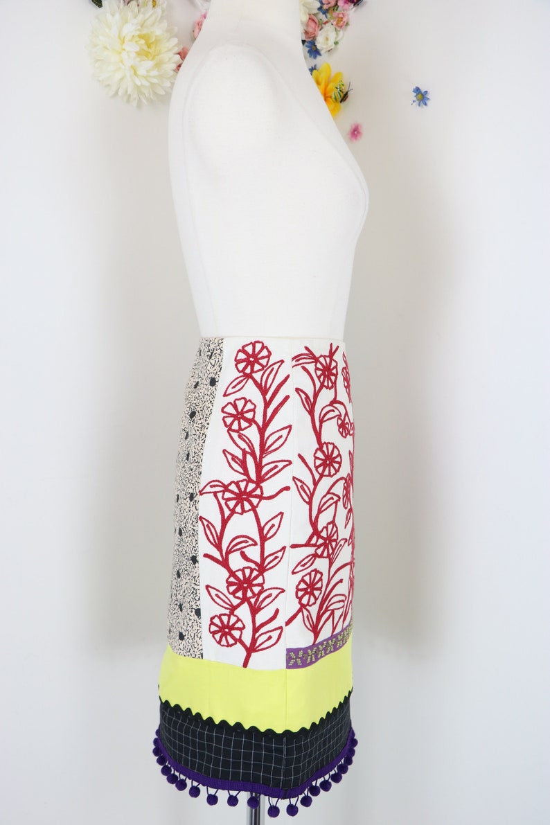 May include: A colorful, patterned skirt with a variety of textures and designs. The skirt features a white background with red floral embroidery, a black and white polka dot panel, and a yellow scalloped hem. The skirt is finished with a black and white checkered panel and purple pom-poms.