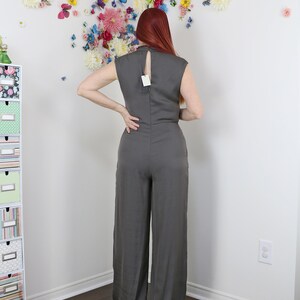 Puede incluir: Mono gris con espalda de ojo de cerradura y perneras anchas. El mono sin mangas tiene un cuello alto y una etiqueta en la espalda. El fondo presenta una pared decorada con flores y mariposas de colores.