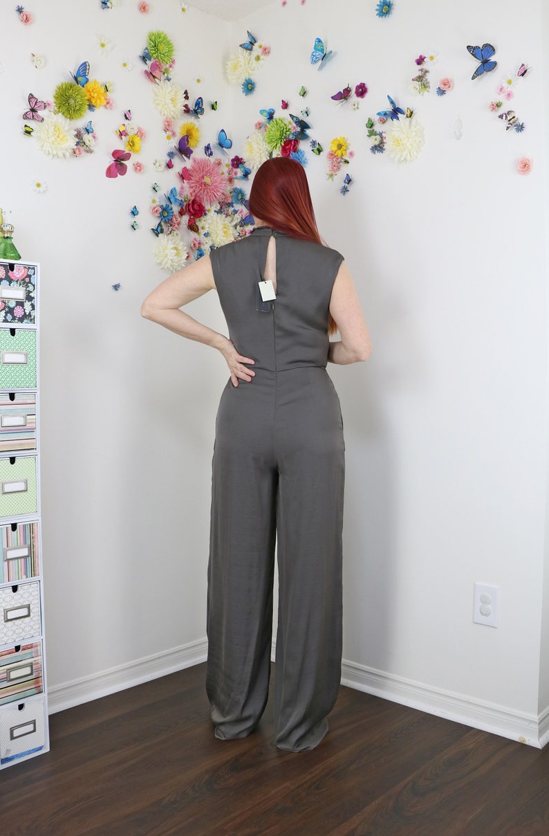 Puede incluir: Mono gris con espalda de ojo de cerradura y perneras anchas. El mono sin mangas tiene un cuello alto y una etiqueta en la espalda. El fondo presenta una pared decorada con flores y mariposas de colores.