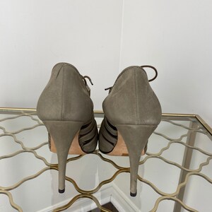 May include: A pair of olive green suede high-heeled shoes. The shoes have a strappy design with thin straps across the foot and a pointed toe. The heels are stiletto style.