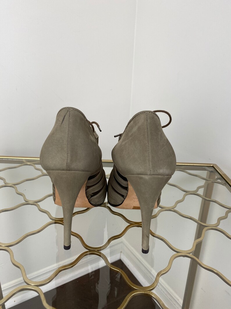 May include: A pair of olive green suede high-heeled shoes. The shoes have a strappy design with thin straps across the foot and a pointed toe. The heels are stiletto style.