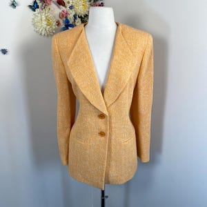May include: A light orange blazer with a textured weave, displayed on a mannequin. The blazer features a notched collar, two brown buttons, and two front pockets. The jacket is a classic style.