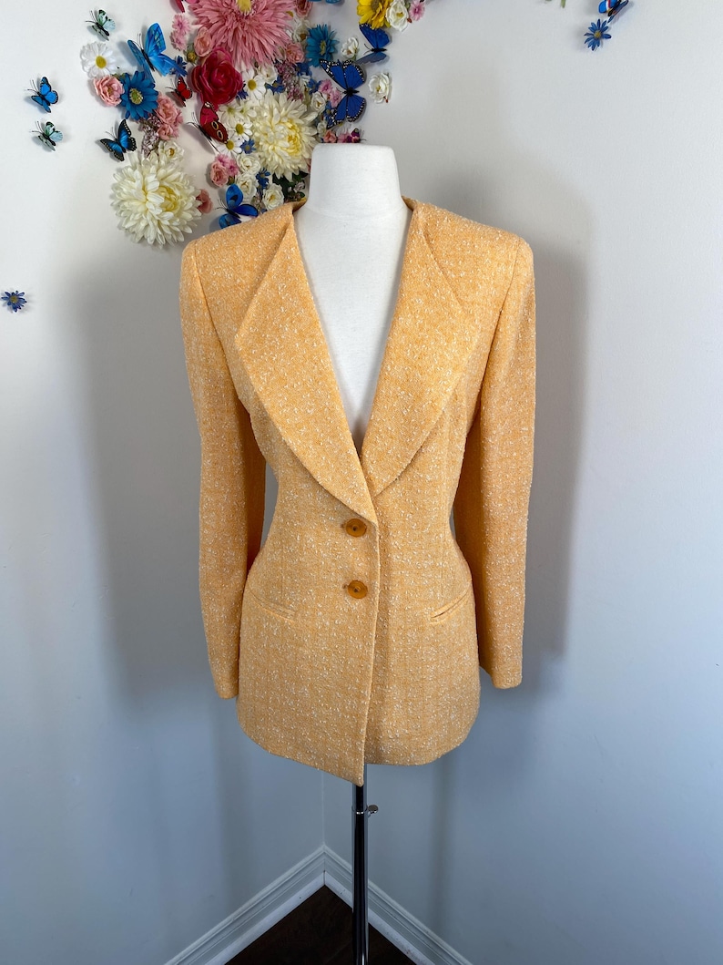 May include: A light orange blazer with a textured weave, displayed on a mannequin. The blazer features a notched collar, two brown buttons, and two front pockets. The jacket is a classic style.