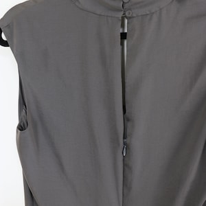 Puede incluir: Vestido gris sin mangas con cuello alto y redondeado y espalda con abertura de cerradura asegurada con dos botones. El vestido tiene cierre de cremallera en la espalda y cuelga de una percha negra.