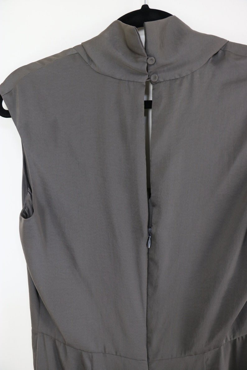Puede incluir: Vestido gris sin mangas con cuello alto y redondeado y espalda con abertura de cerradura asegurada con dos botones. El vestido tiene cierre de cremallera en la espalda y cuelga de una percha negra.