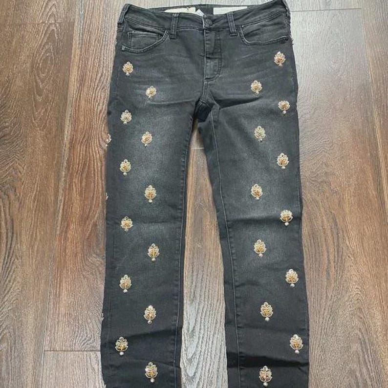 May include: Dark grey denim jeans with a slim fit, featuring gold and cream-coloured embroidered floral patterns. The jeans have a classic five-pocket design and a button and zip closure. The embroidery is evenly spaced across the front of the jeans.