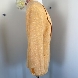 May include: A light orange blazer with a textured weave. The blazer has a notched lapel, two front pockets, and three buttons on each cuff. The blazer is displayed on a mannequin.