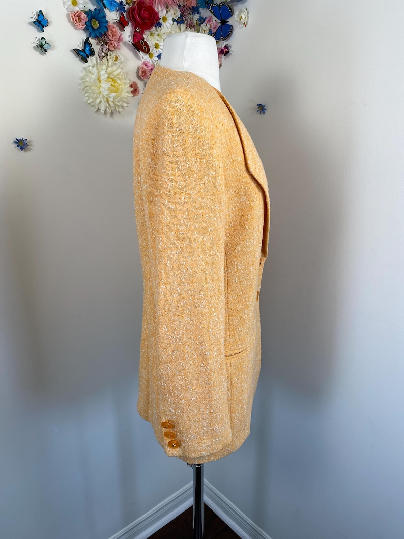 May include: A light orange blazer with a textured weave. The blazer has a notched lapel, two front pockets, and three buttons on each cuff. The blazer is displayed on a mannequin.