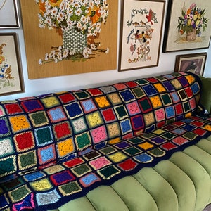 May include: A vibrant, hand-crocheted blanket with a patchwork design, resting on a green sofa. The blanket displays a variety of colours, such as red, blue, yellow, and green. Framed wall art with floral and bird motifs is visible in the background.