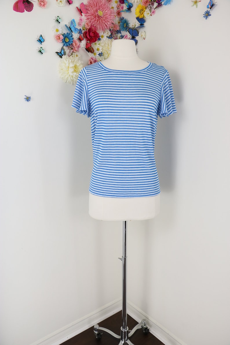 May include: A blue and white striped t-shirt with short sleeves is displayed on a mannequin. The shirt has a round neckline and a classic horizontal stripe pattern. The mannequin is on a silver stand, and the background features floral decorations.