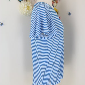 May include: A light blue and white striped top with short sleeves. The shirt is displayed on a mannequin. The top has a V-neck and a split at the bottom hem. The background features floral and butterfly decorations.
