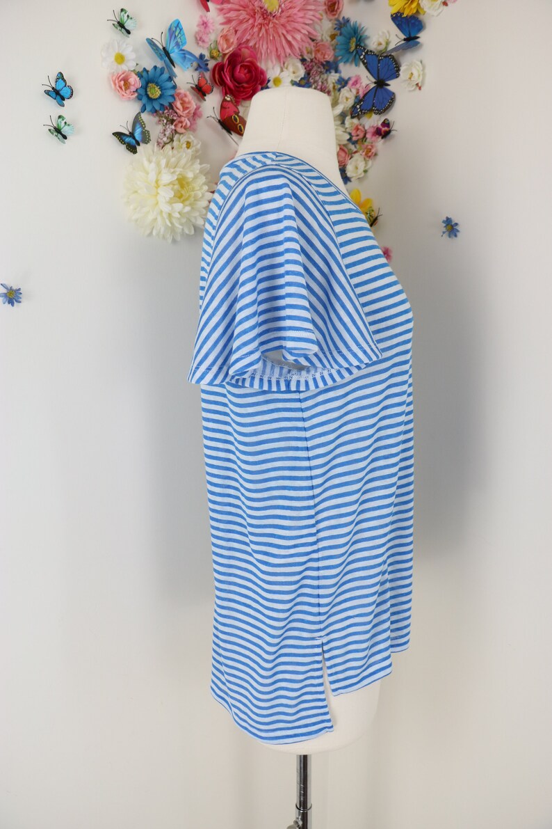 May include: A light blue and white striped top with short sleeves. The shirt is displayed on a mannequin. The top has a V-neck and a split at the bottom hem. The background features floral and butterfly decorations.