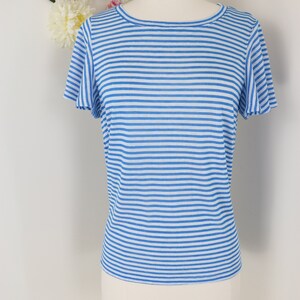 May include: A blue and white striped short-sleeve shirt displayed on a mannequin. The shirt has a round neckline and a relaxed fit. The background features a white wall decorated with artificial flowers and butterflies.