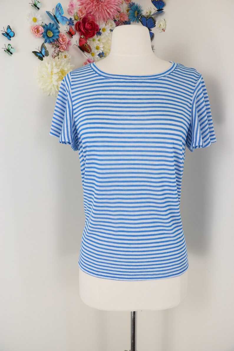 May include: A blue and white striped short-sleeve shirt displayed on a mannequin. The shirt has a round neckline and a relaxed fit. The background features a white wall decorated with artificial flowers and butterflies.