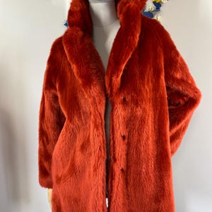 May include: A vibrant orange faux fur coat with a wide collar and long sleeves. The coat has a button closure and is displayed on a mannequin. The coat's plush texture and bold color make it a statement piece.