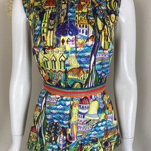 May include: A sleeveless romper with a vibrant, artistic print depicting buildings, trees, and water in yellow, blue, and green hues. The romper has a round neckline and is cinched at the waist with a decorative belt.