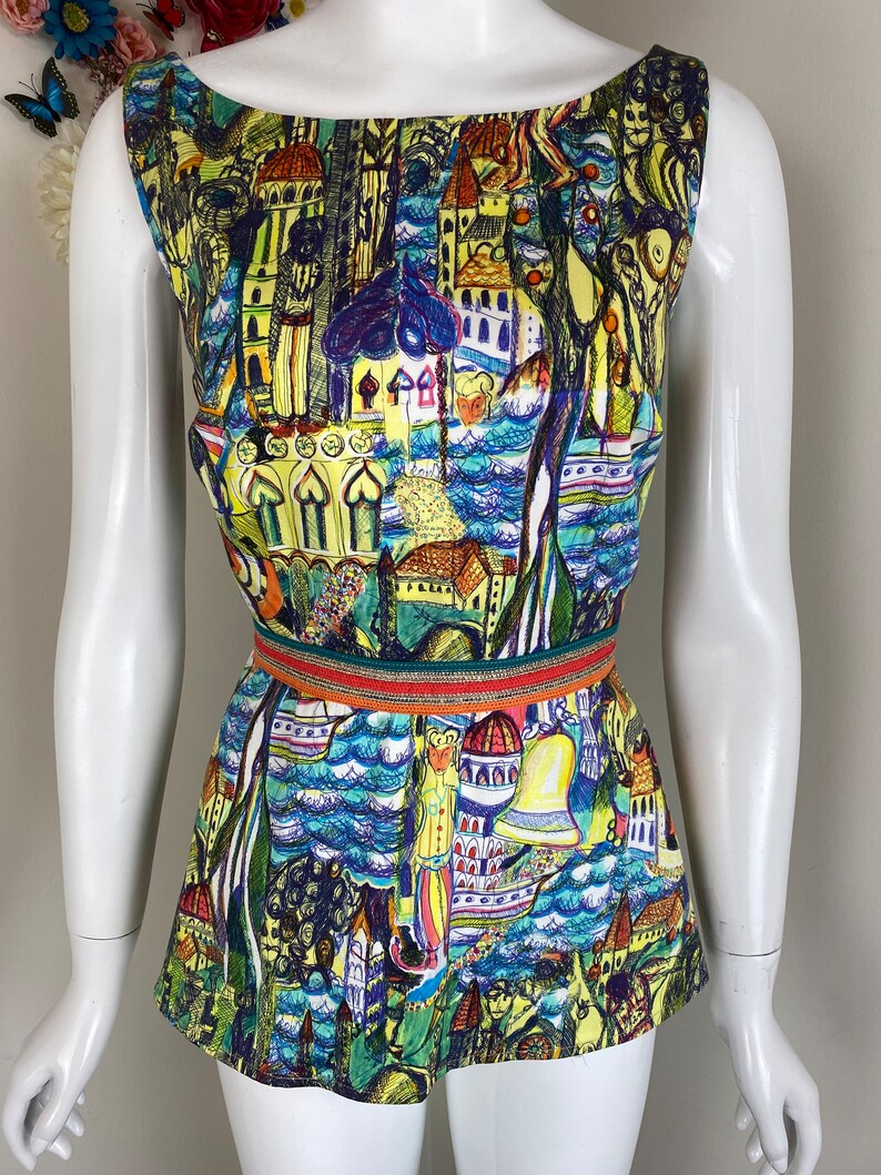 May include: A sleeveless romper with a vibrant, artistic print depicting buildings, trees, and water in yellow, blue, and green hues. The romper has a round neckline and is cinched at the waist with a decorative belt.