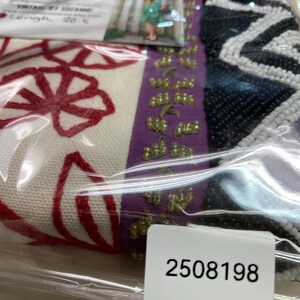 May include: A close-up of a vintage textile, featuring red floral embroidery on a cream background. A purple ribbon with gold embellishments and a black and white beaded section are visible. A tag with the number 2508198 is also present.
