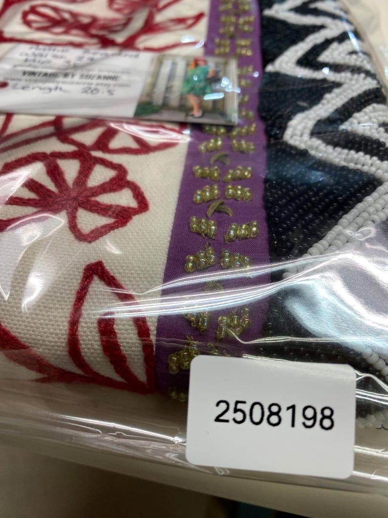 May include: A close-up of a vintage textile, featuring red floral embroidery on a cream background. A purple ribbon with gold embellishments and a black and white beaded section are visible. A tag with the number 2508198 is also present.