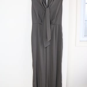 Puede incluir: Mono sin mangas gris carb&oacute;n colgado de una percha negra. El mono presenta un cuello drapeado con un largo detalle de corbata. La prenda tiene un corpi&ntilde;o ajustado y pantalones de pernera ancha, creando una silueta elegante y moderna.