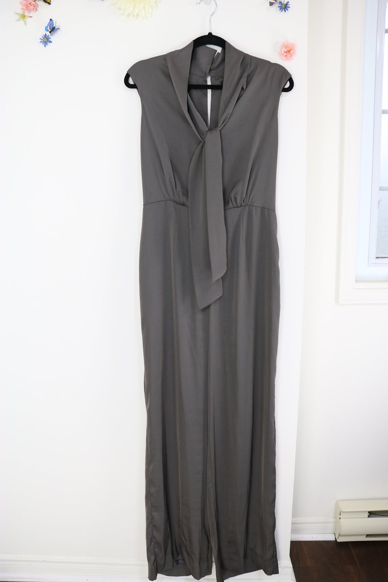 Puede incluir: Mono sin mangas gris carb&oacute;n colgado de una percha negra. El mono presenta un cuello drapeado con un largo detalle de corbata. La prenda tiene un corpi&ntilde;o ajustado y pantalones de pernera ancha, creando una silueta elegante y moderna.