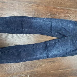 May include: Dark wash denim jeans, lying flat on a wooden surface. The jeans feature a classic five-pocket design and a straight leg cut. The denim has subtle fading and whiskering details.