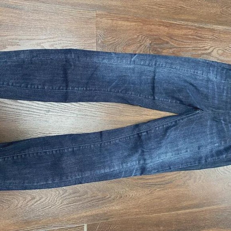 May include: Dark wash denim jeans, lying flat on a wooden surface. The jeans feature a classic five-pocket design and a straight leg cut. The denim has subtle fading and whiskering details.