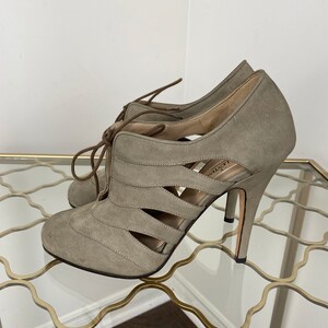 May include: A pair of gray suede high-heeled shoes with cut-out detailing. The shoes have a lace-up design and a stiletto heel. The heels are approximately 4 inches (10 cm) high. The shoes are displayed on a glass surface.