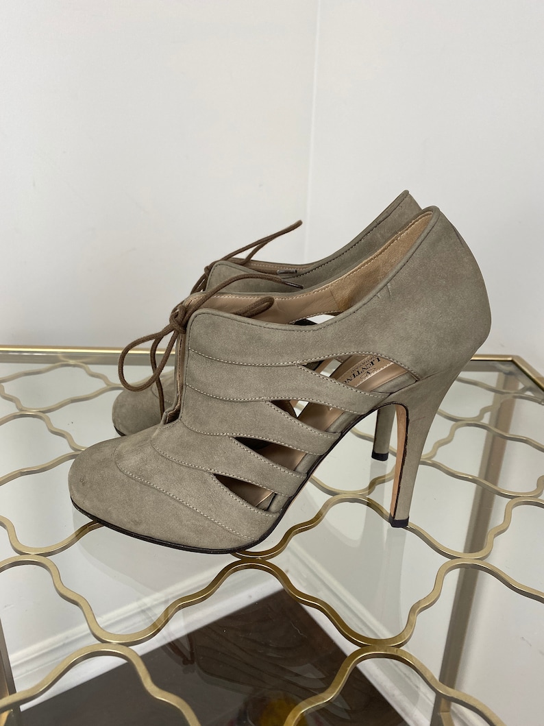 May include: A pair of gray suede high-heeled shoes with cut-out detailing. The shoes have a lace-up design and a stiletto heel. The heels are approximately 4 inches (10 cm) high. The shoes are displayed on a glass surface.