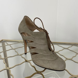 May include: A taupe suede high-heeled shoe with cut-out detailing and a lace-up closure. The shoe features a stiletto heel and a rounded toe. The design includes horizontal cut-out accents and a brown lace. The shoe is displayed on a glass surface.