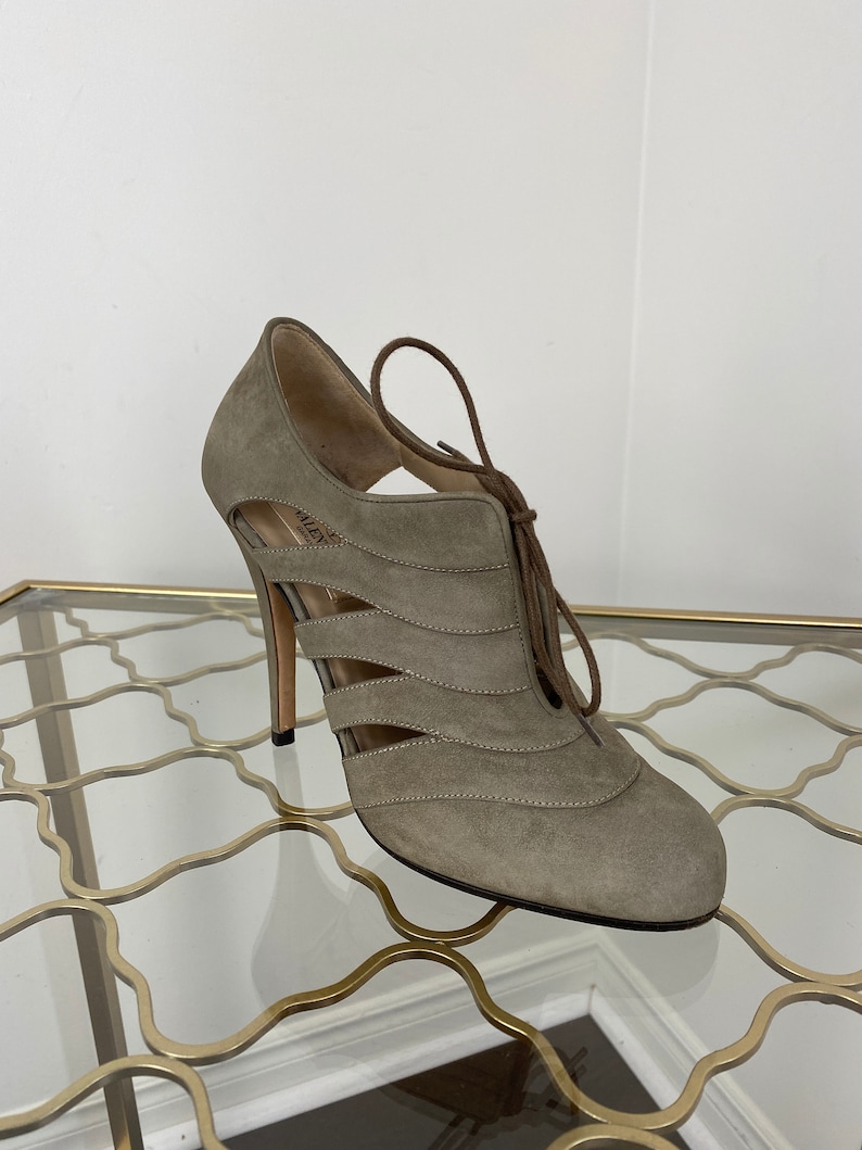 May include: A taupe suede high-heeled shoe with cut-out detailing and a lace-up closure. The shoe features a stiletto heel and a rounded toe. The design includes horizontal cut-out accents and a brown lace. The shoe is displayed on a glass surface.