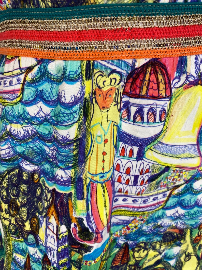 May include: Close-up of a vibrant fabric with a detailed illustration. The design features a whimsical cityscape with figures, buildings, and water elements in shades of blue, yellow, and red. The top edge has a decorative trim with green, orange, and gold accents.