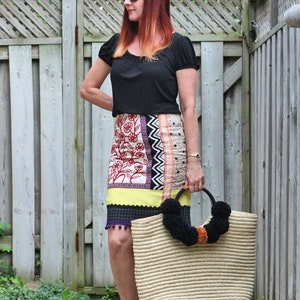 May include: A woman wearing a black top and a patterned skirt, holding a large straw tote bag with black pom-poms. The skirt features floral and geometric designs. She is wearing black sandals.