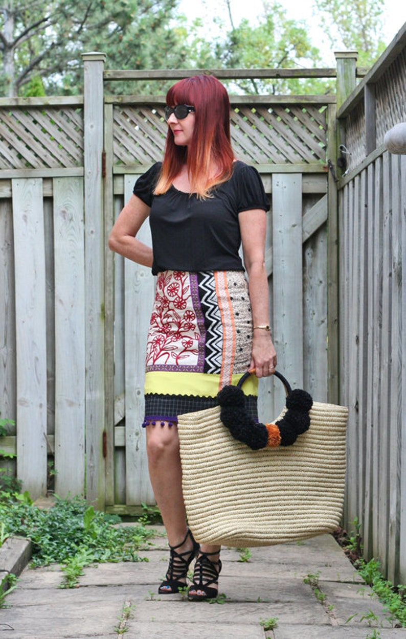 May include: A woman wearing a black top and a patterned skirt, holding a large straw tote bag with black pom-poms. The skirt features floral and geometric designs. She is wearing black sandals.