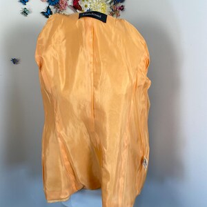 May include: A vintage orange silk jacket interior, showcasing the lining and construction. The label reads "Louis F&eacute;raud". The jacket is displayed on a mannequin, with a floral arrangement in the background. The silk has a subtle sheen.