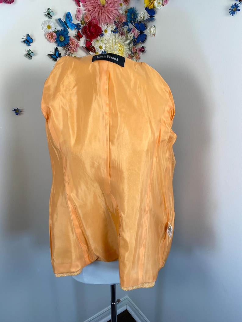 May include: A vintage orange silk jacket interior, showcasing the lining and construction. The label reads "Louis F&eacute;raud". The jacket is displayed on a mannequin, with a floral arrangement in the background. The silk has a subtle sheen.