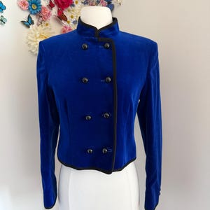 May include: A vibrant royal blue velvet jacket featuring a high mandarin collar, long sleeves, and a double-breasted front closure with eight black buttons. The jacket is finished with black piping along the edges, collar, and front opening.