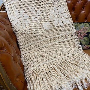 May include: A cream-colored crochet throw blanket featuring an intricate openwork grid pattern, floral motifs, and a decorative border with long, dangling fringe tassels along the bottom edge. The textile has a textured, knotted appearance.