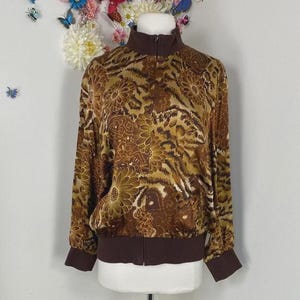 May include: A brown zip-up jacket with a floral and animal print design. The jacket has a high collar, long sleeves, and ribbed cuffs and hem. The color palette includes shades of brown, tan, and olive green.