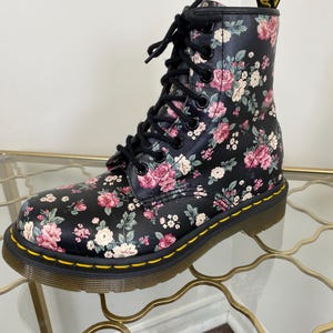 May include: A black ankle boot featuring a floral pattern with pink, white, and green flowers. The boot has black laces, black eyelets, a yellow welt stitch along the sole, and a yellow and black heel pull tab with the text AirWair.