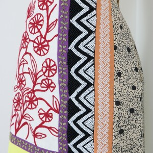 May include: A colorful skirt with a patchwork design. The skirt features panels of white fabric with red floral embroidery, black beaded zigzags, and a tan fabric with black polka dots. A yellow border with a black and white checkered trim completes the design.