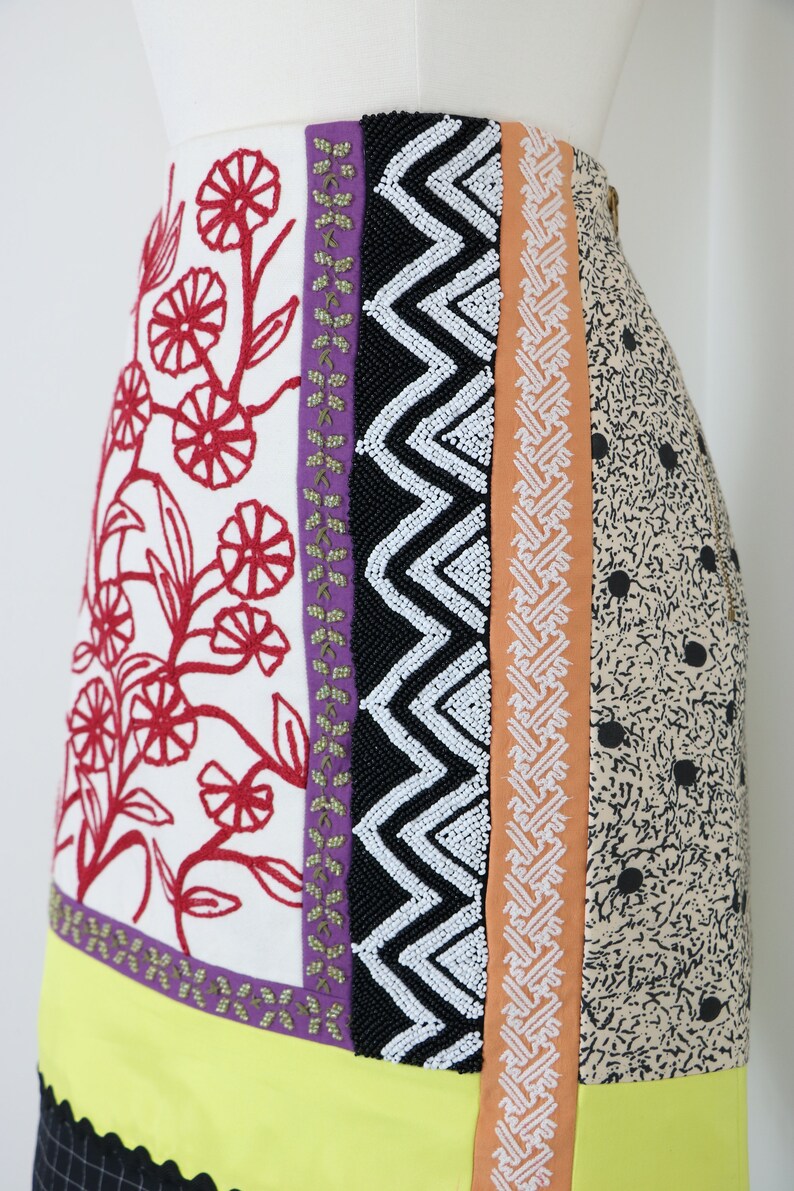 May include: A colorful skirt with a patchwork design. The skirt features panels of white fabric with red floral embroidery, black beaded zigzags, and a tan fabric with black polka dots. A yellow border with a black and white checkered trim completes the design.