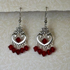 Puede incluir: Un par de pendientes colgantes plateados con un diseño en forma de corazón. Cada pendiente presenta una cuenta central roja en forma de diamante y múltiples cuentas rojas colgantes, creando un efecto en capas. Accesorio decorativo.