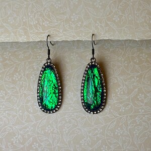 May include: A pair of teardrop-shaped earrings with a vibrant green, iridescent center. The earrings are framed with a dark metal and surrounded by small, clear crystals. The hooks are also dark metal. The background is a cream color with a floral pattern.
