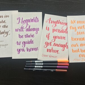 May include: Four white cards with inspirational quotes from the Harry Potter series written in calligraphy. The quotes are: "When in doubt, go to the library" by Hermione Granger, "Hogwarts will always be there to guide you home", "Anything is possible if you've got enough nerve." by Ginny Weasley, and "We must try not to sink beneath our anguish, but battle on." by Albus Dumbledore.