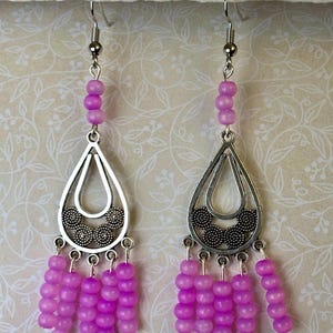 May include: A pair of dangle earrings featuring silver-toned teardrop-shaped pendants with a floral design. The earrings have multiple strands of small, vibrant purple beads hanging from the bottom and a few beads at the top.