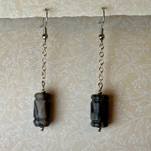 Puede incluir: Un par de pendientes colgantes con cuentas cilíndricas de piedra gris y negra. Las cuentas cuelgan de cadenas y ganchos plateados. Los pendientes se exhiben sobre un fondo crema con un estampado floral.