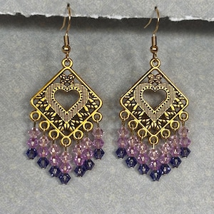 Puede incluir: Un par de pendientes colgantes dorados. Cada pendiente presenta un diseño de filigrana en forma de diamante con un corazón recortado y cuentas colgantes en tonos morados y azules. Cierre de gancho.