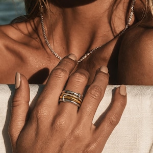 May include: A stack of four rings worn on a finger, featuring two smooth silver-toned bands, one gold-toned band, and one band encrusted with small clear stones. A delicate silver-toned chain necklace with small clear stones rests around the neck.