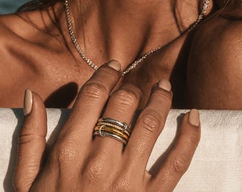 May include: A stack of four rings worn on a finger, featuring two smooth silver-toned bands, one gold-toned band, and one band encrusted with small clear stones. A delicate silver-toned chain necklace with small clear stones rests around the neck.